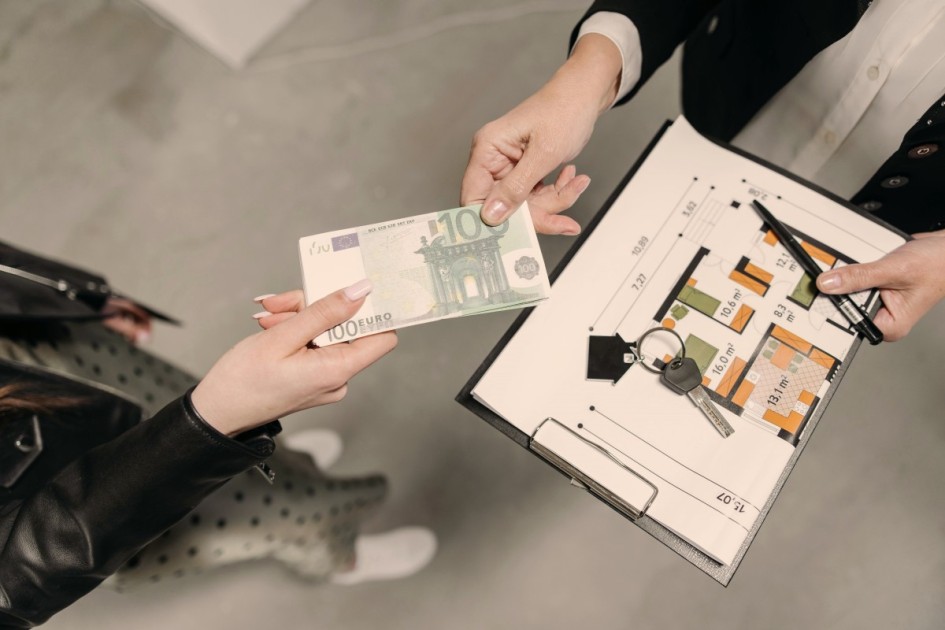 a birds eye view of a real estate agent holding a clipboard of a housing floorplan and keys in one hand accepts a hundred dollar bill (signifying a lowball offer) from a person in a long dress in the other hand 